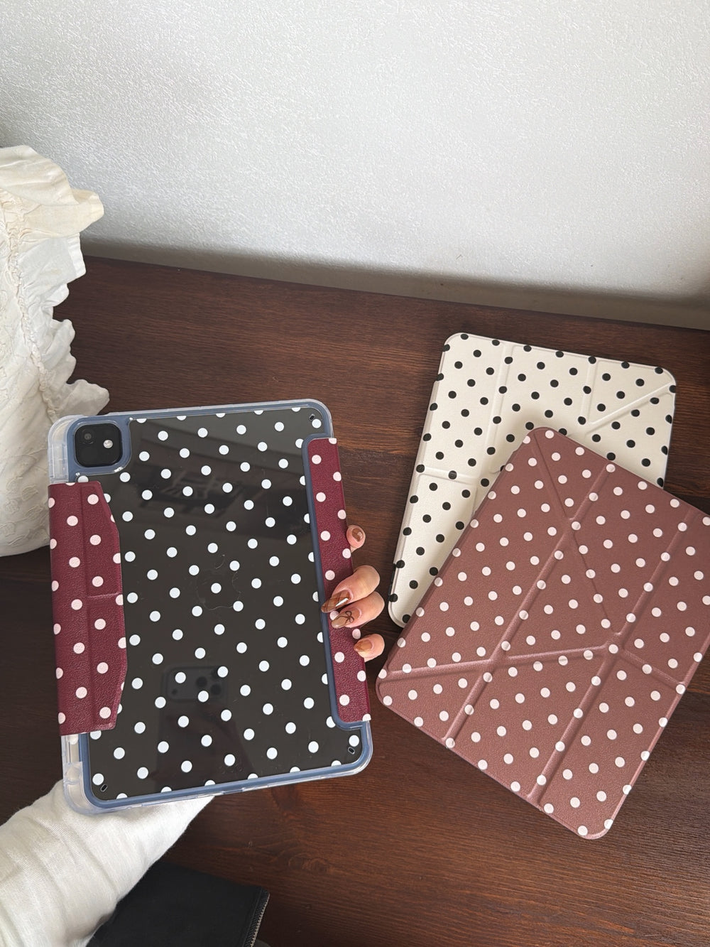 A flat lay showcasing an aesthetic polka dot iPad case collection in burgundy, white, and black-clear variations on a dark wood desk.