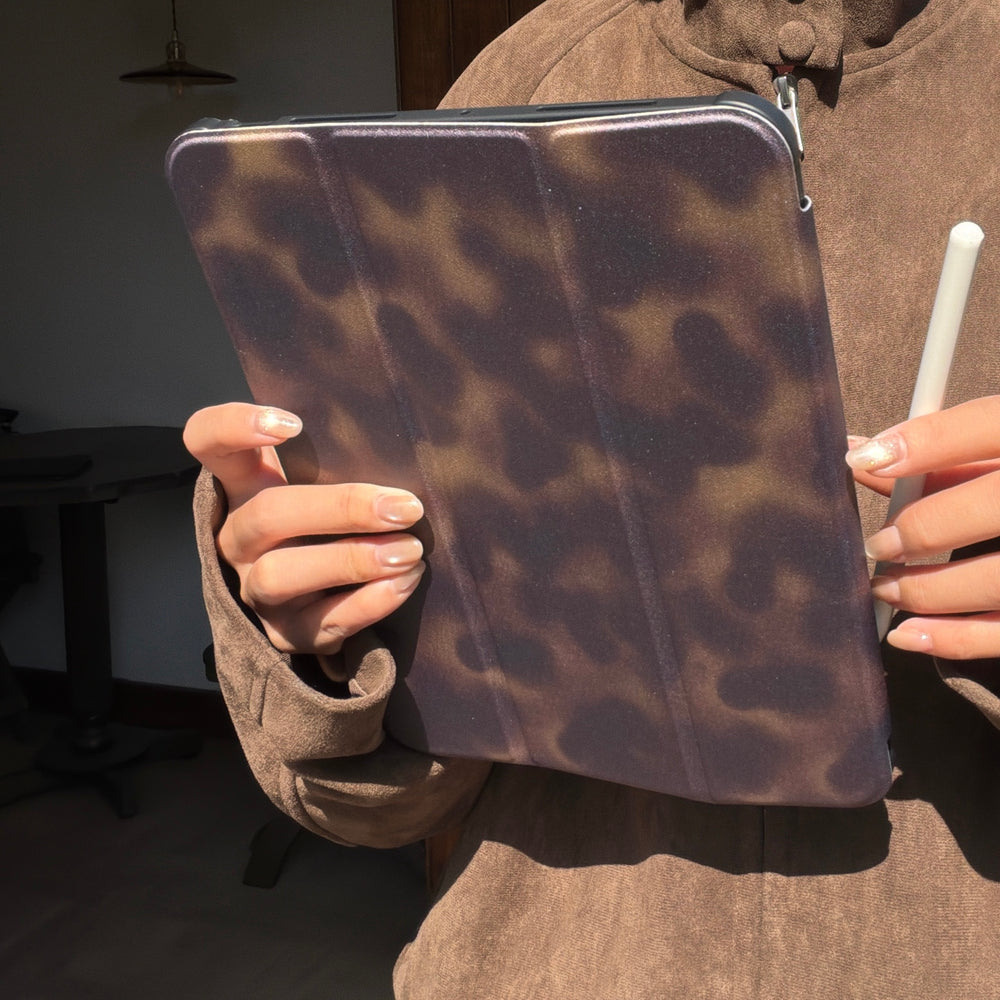 Close-up of a person wearing a brown suede jacket holding a closed aesthetic tortoiseshell ipad case while gripping a white Apple Pencil.