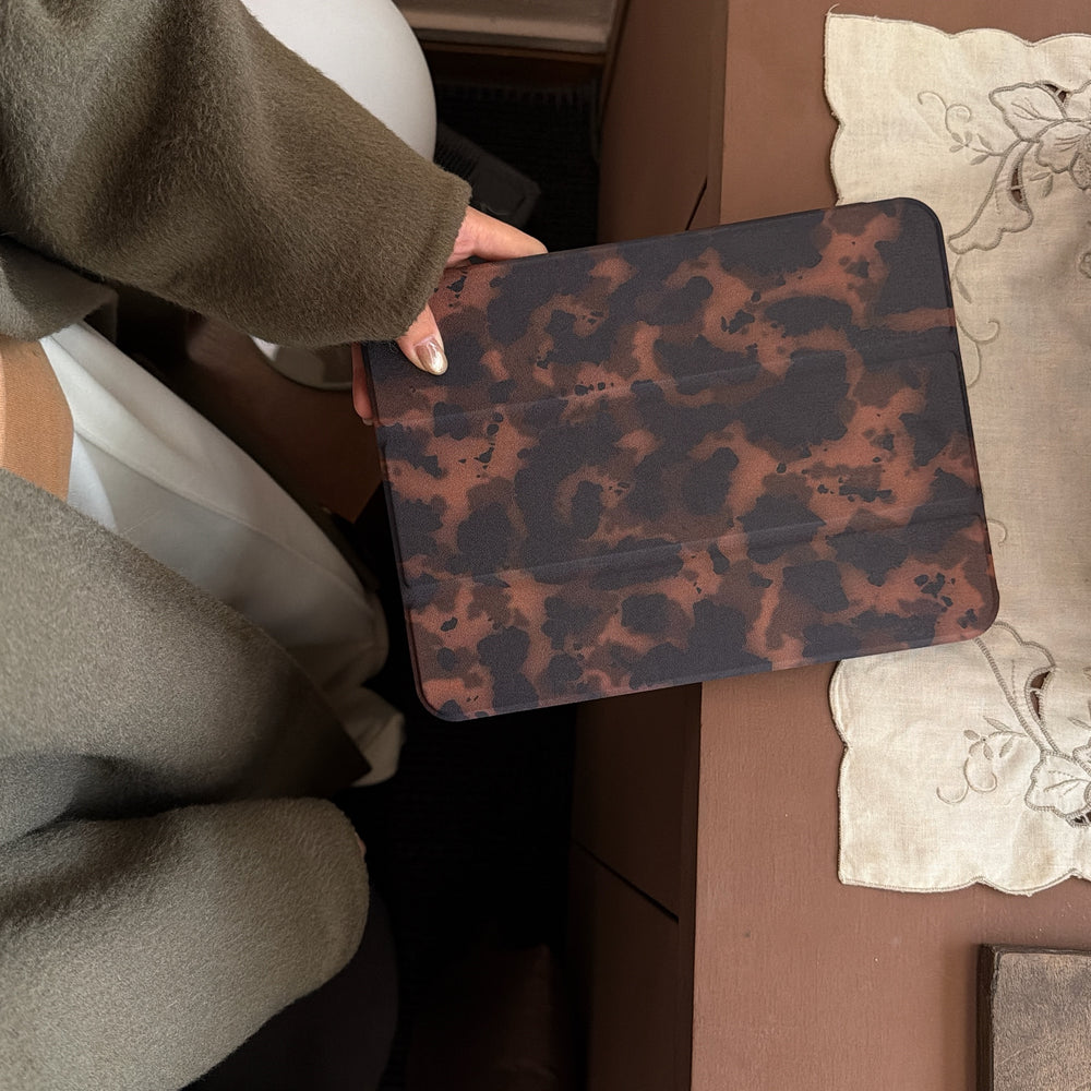 A person wearing an olive green wool coat holding a closed tortoiseshell tri fold tablet case over a brown desk with a vintage embroidered white lace mat.