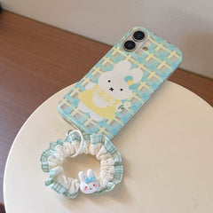 A blue plaid cartoon bunny phone case resting on a white stand with its matching green ruffled scrunchie wristlet.