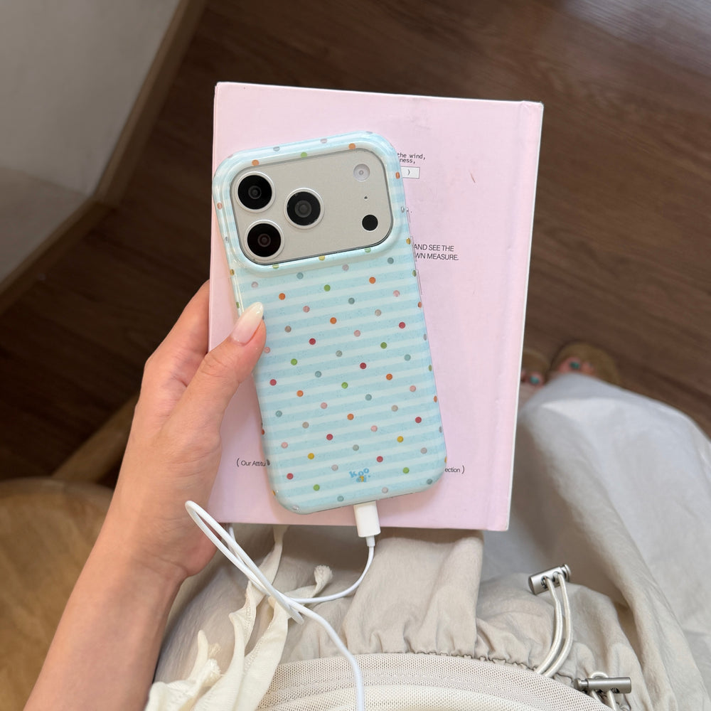 A hand holding a blue stripe polka dot iPhone case while plugged into a white charger over a pink book.