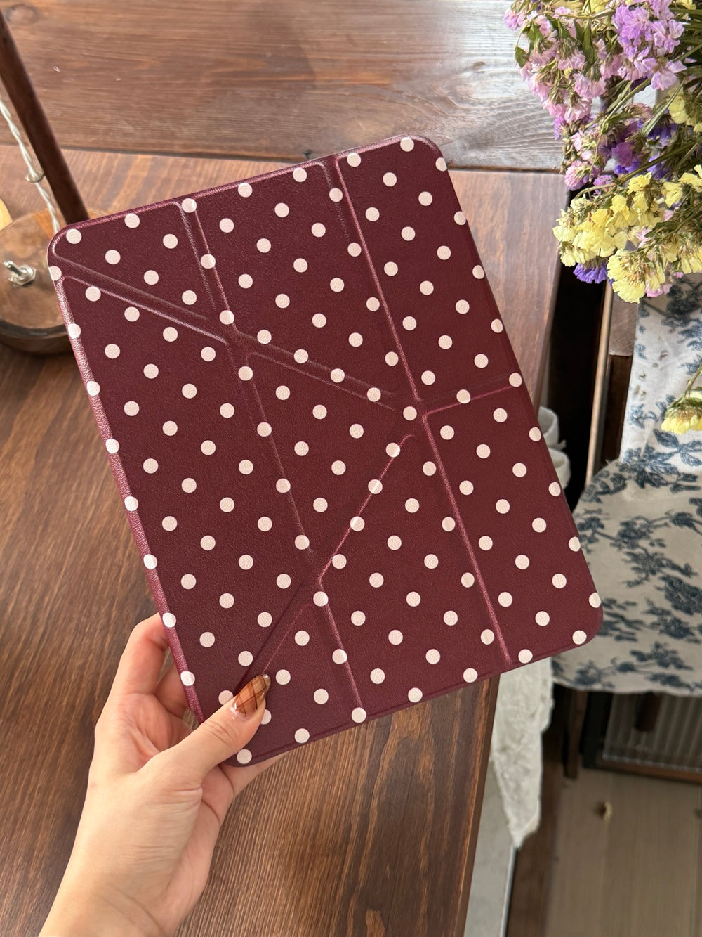 A hand holding a closed burgundy polka dot iPad case with origami geometric folding lines over a wooden table.