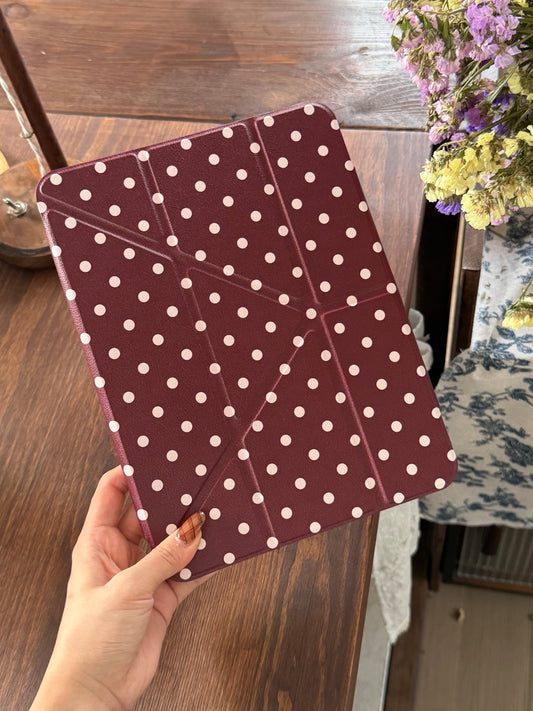 A hand holding a closed burgundy polka dot iPad case with origami geometric folding lines over a wooden table.