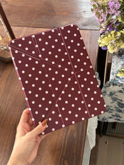 A hand holding a closed burgundy polka dot iPad case with origami geometric folding lines over a wooden table.