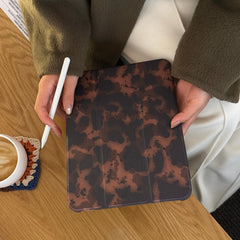 Top-down lifestyle shot of a chic animal print ipad case held with a white stylus on a light wooden table, next to a coffee cup on a colorful crocheted coaster.