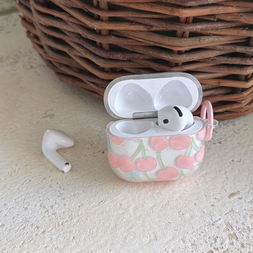 An open coquette cherry AirPods case displaying a white earbud next to a rustic wicker basket.