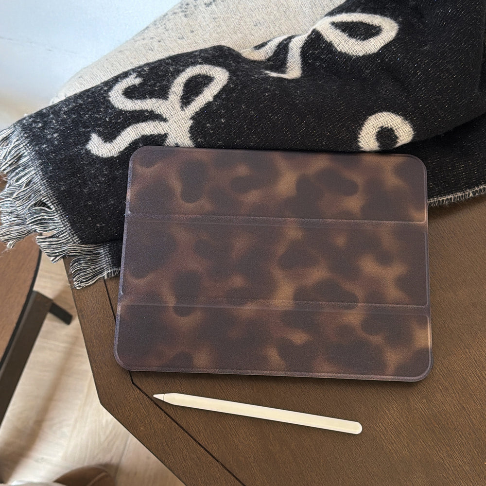 An aesthetic tortoiseshell ipad case lying flat on a wooden desk next to a white Apple Pencil and a black and white patterned bow blanket for a cozy workspace.