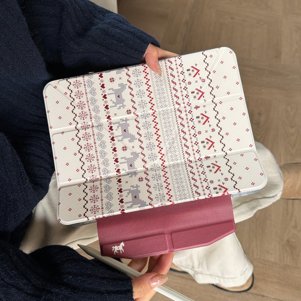 A top-down view of a closed cozy cabin core iPad folio on someone's lap, emphasizing the wine-red magnetic clasp with a white pixel horse icon.