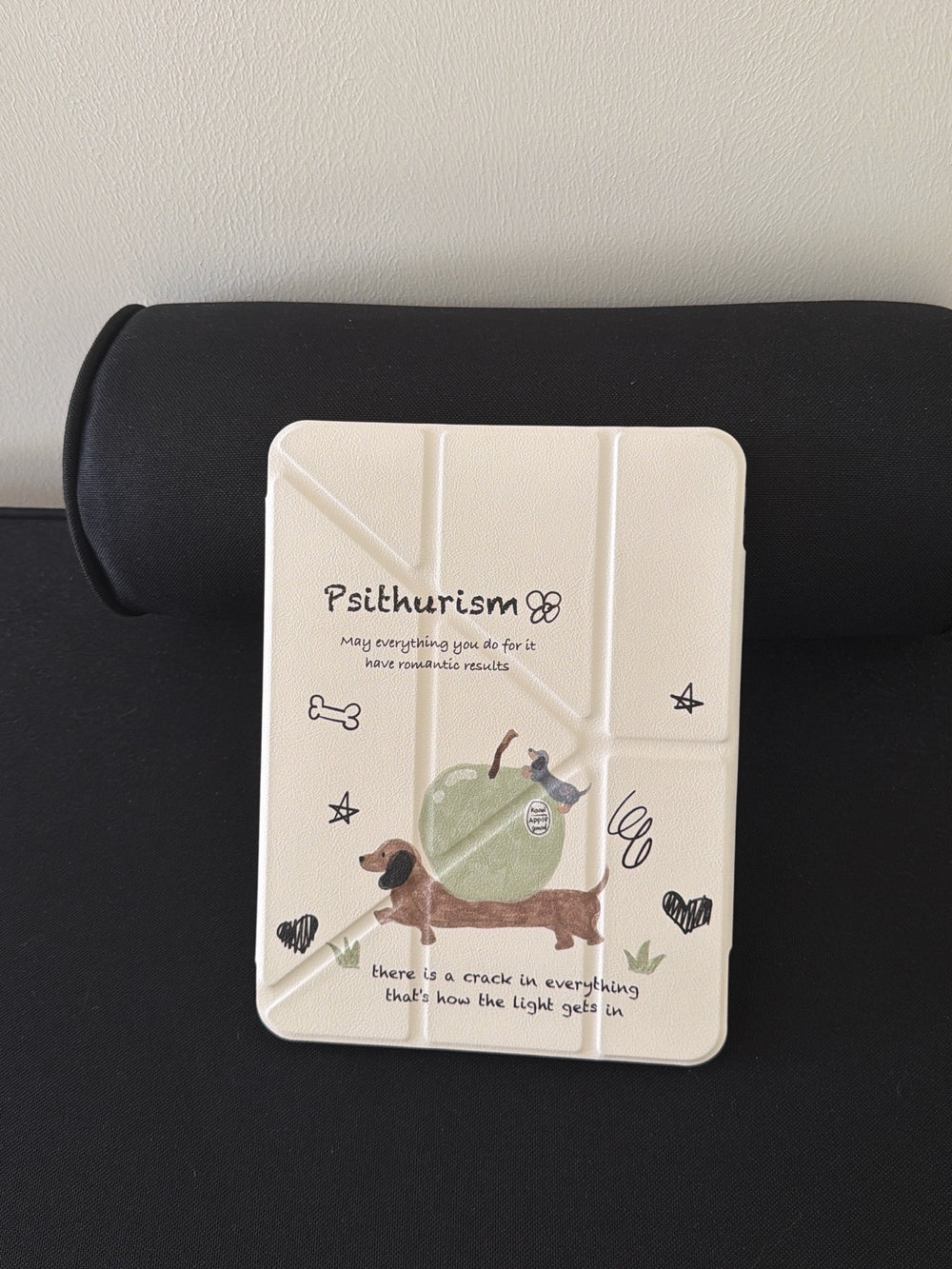 The closed front view of a cute dachshund ipad case standing vertically against a black bolster pillow, highlighting the cream origami cover with hand-drawn dog and apple doodles.