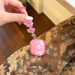 A hand lifting a pink hibiscus flower AirPods case by its round pink carabiner hook over a brown floral backdrop.