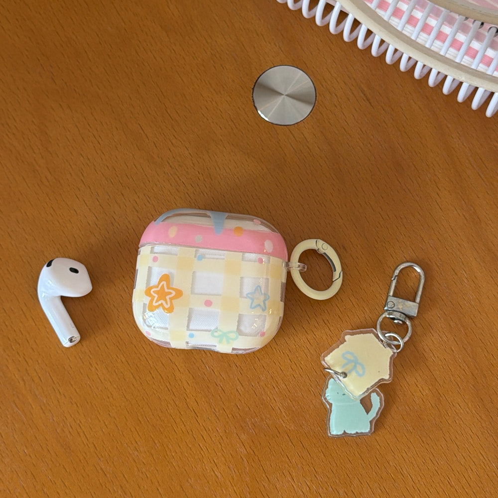A flatlay of a pastel pink drip AirPods case on a wooden desk next to a single wireless earbud and a cat charm.
