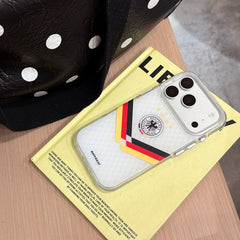 A silver Germany 4-stars mirror phone case lying flat on a yellow book next to a polka dot bag.