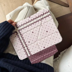A closed heart lace ribbon pink iPad smart cover resting on a model's lap, emphasizing the textured geometric folding lines and dark pink clasp.