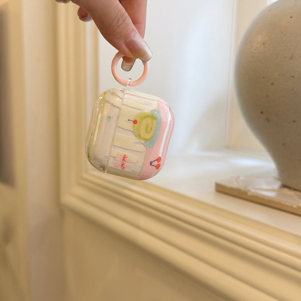 A hand dangling a kawaii cake design AirPods case by its pastel pink circular carabiner ring hook.