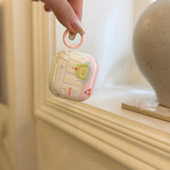 A hand dangling a kawaii cake design AirPods case by its pastel pink circular carabiner ring hook.