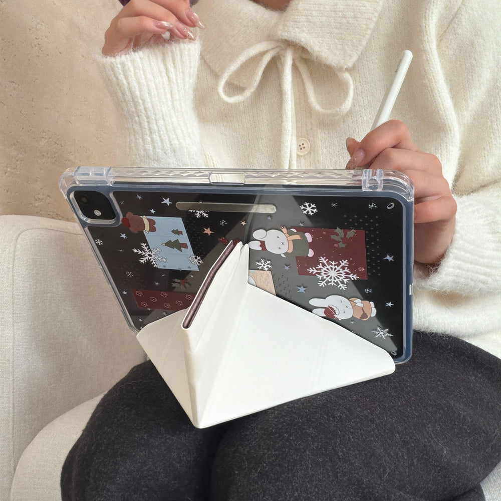 A person holding an iPad in a vertical position supported by a white magnetic origami stand, showing the clear back panel printed with cute winter Miffy bunny graphics.
