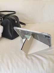 A kawaii origami tablet cover propped up horizontally on a cream-white fabric sofa next to a black leather handbag, utilizing its geometric folding stand.