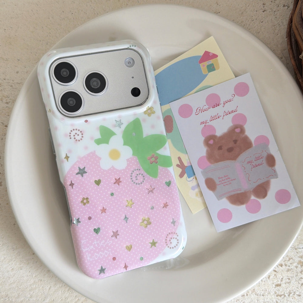 Top view of a kawaii pink strawberry iPhone case with a white camera frame resting on a ceramic plate next to a cartoon bear postcard.