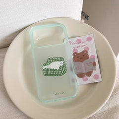 A kawaii clear white cat on a green couch iPhone case shell resting on a ceramic plate with a cartoon bear postcard.