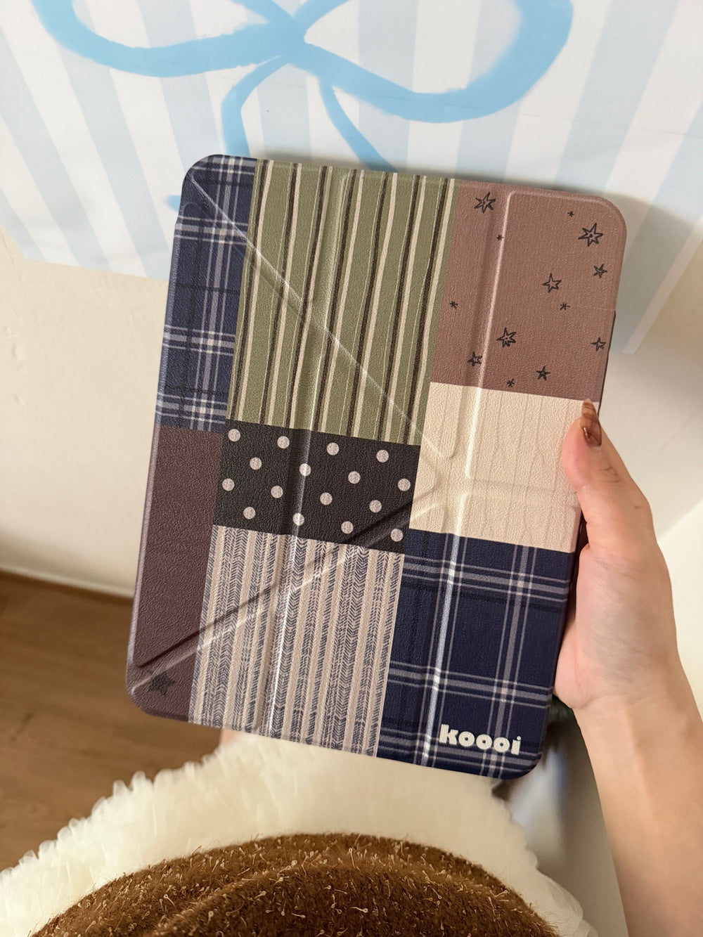 An angled first-person view of a hand holding the closed Koooi vintage patchwork plaid smart cover, detailing the rustic textures of plaid, grids, and polka dots.