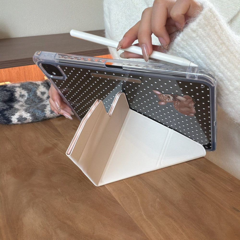 An iPad supported in landscape mode by a cream-colored origami viewing stand on a wooden desk, while a hand holds a white stylus pencil over it.