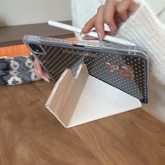 An iPad supported in landscape mode by a cream-colored origami viewing stand on a wooden desk, while a hand holds a white stylus pencil over it.