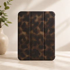 A luxury tortoiseshell ipad case standing vertically on a minimalist beige countertop next to a white ceramic vase with green leaves under soft casting shadows.