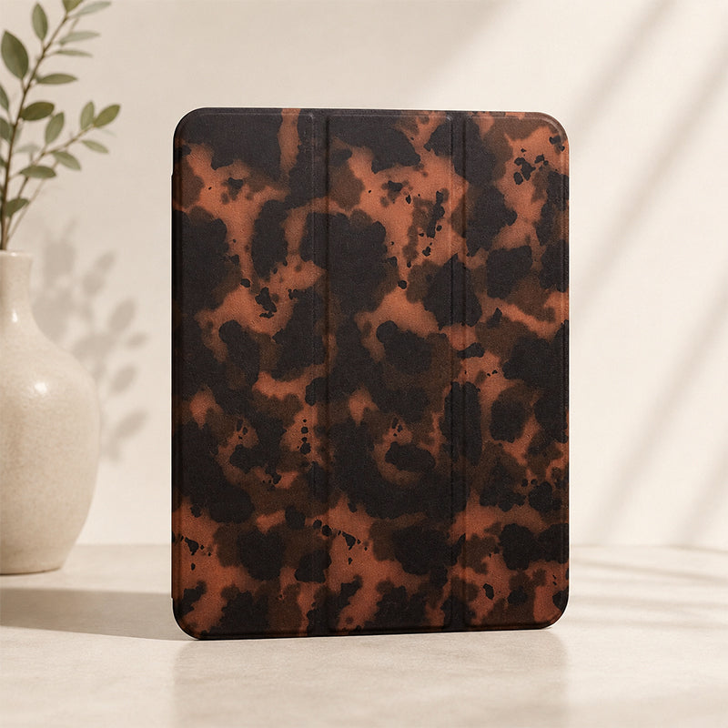 A luxury tortoiseshell ipad case standing vertically on a minimalist beige countertop under soft aesthetic shadows, next to a white ceramic vase with green leaves.