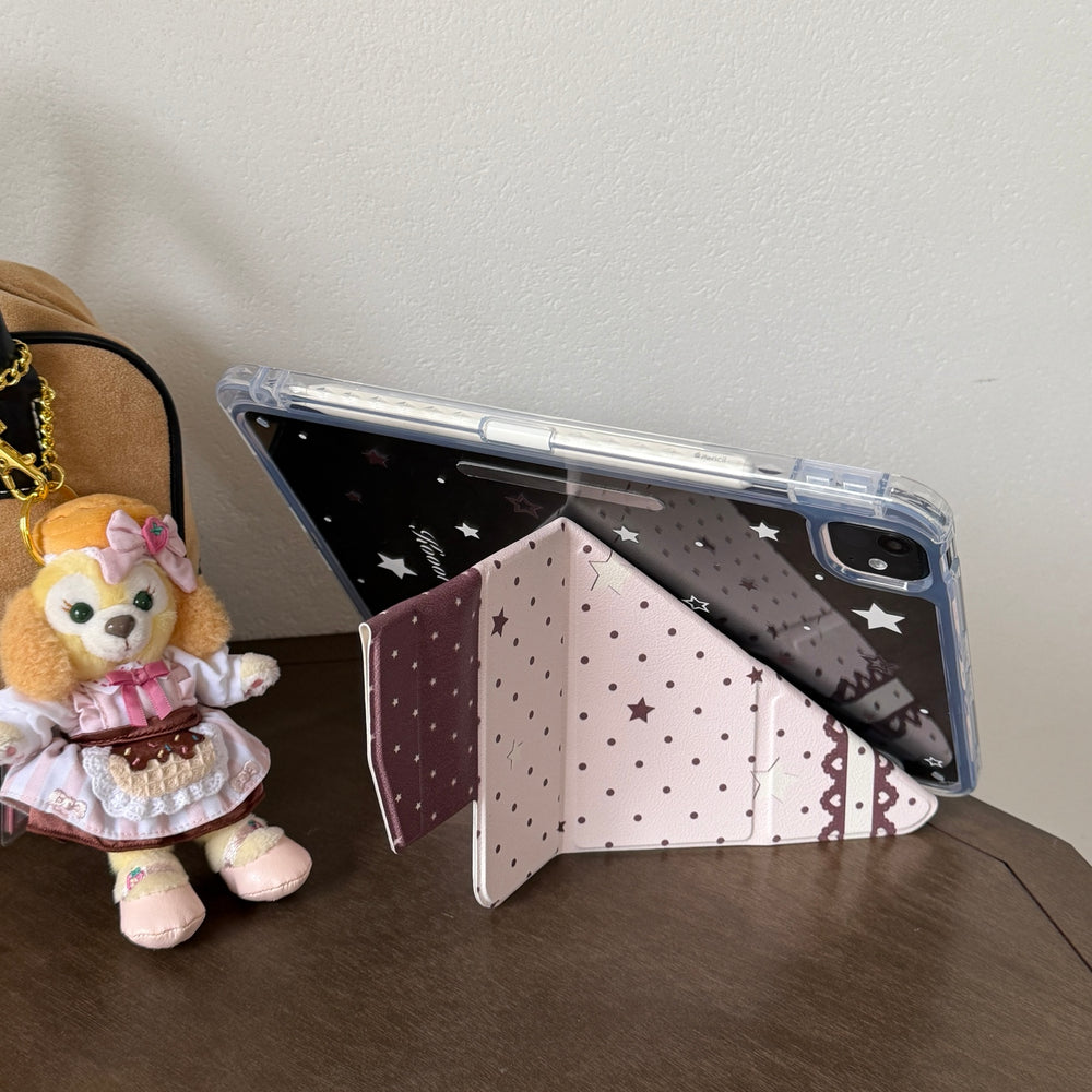 An iPad supported in landscape mode by a pink origami folding stand next to a cute plush toy on a dark wooden table.