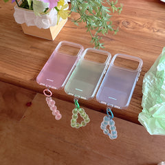 Three pastel-colored clear bumper phone cases in pink, green, and blue lined up on a wooden table with matching straps.