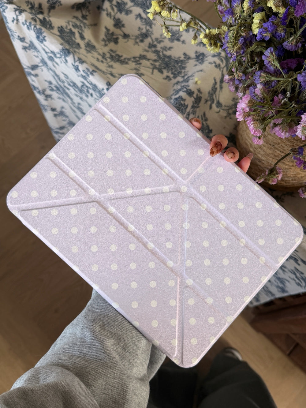A hand holding a pastel lavender polka dot ipad cover with structural origami fold lines next to a basket of dried purple flowers.