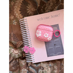 A translucent pink hibiscus AirPods case with a pink ring loop resting on top of a pink striped notebook.