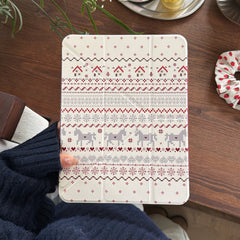 A hand holding a scandinavian pattern iPad smart cover showing pixel art houses and horses over a dark wooden desktop next to a strawberry scrunchie.
