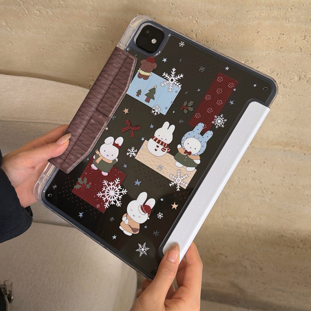Hands holding the printed clear back shell of a shockproof Miffy iPad case, displaying the dark transparent panel with adorable winter bunny graphics and a brown magnetic clasp.