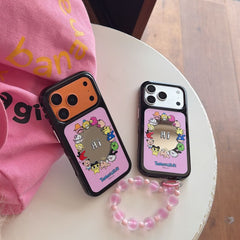 Two Tamagotchi mirror iPhone cases in silver and orange colors on a white tabletop with a pink beaded strap.