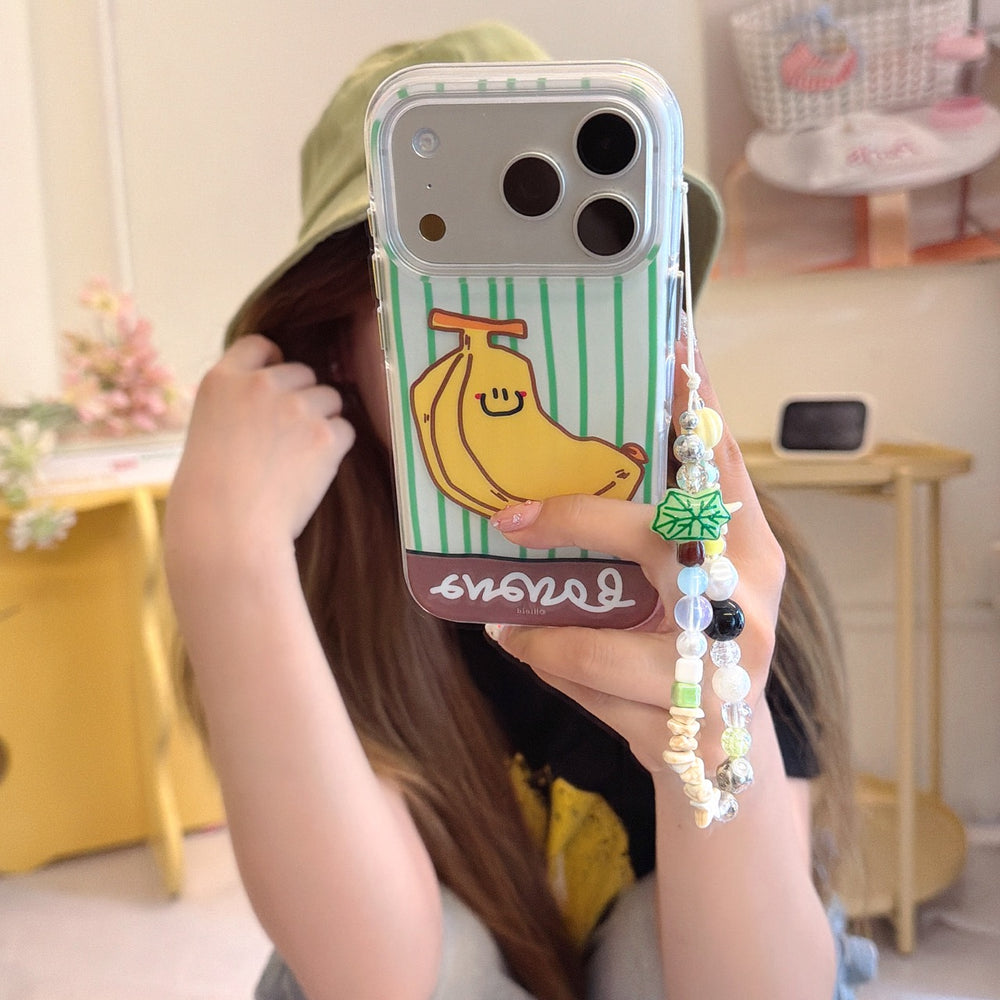 A mirror selfie of a girl wearing a green bucket hat, showing off her cute y2k-style striped banana phone case with a beaded strap.
