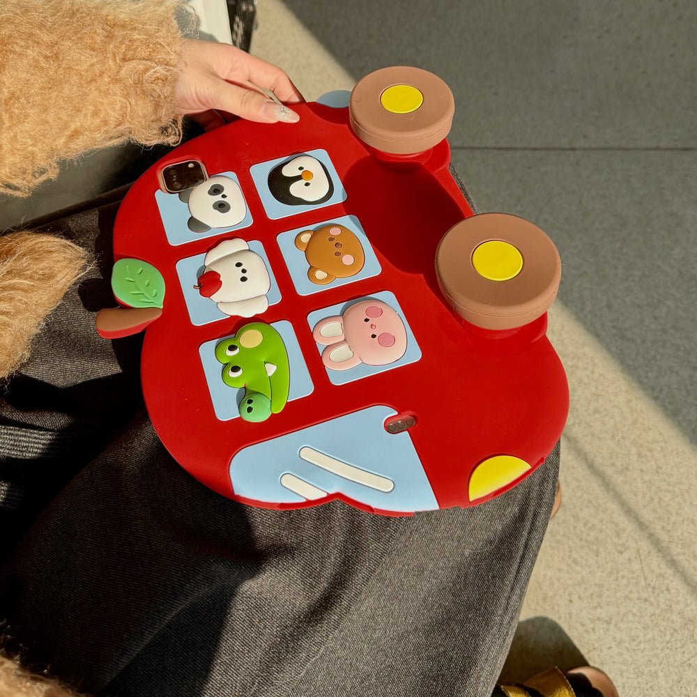 Top-down view of the red apple bus iPad cover resting on a lap, showcasing six 3D cartoon animals in blue windows, a green leaf stem, and two large brown wheels with yellow centers.