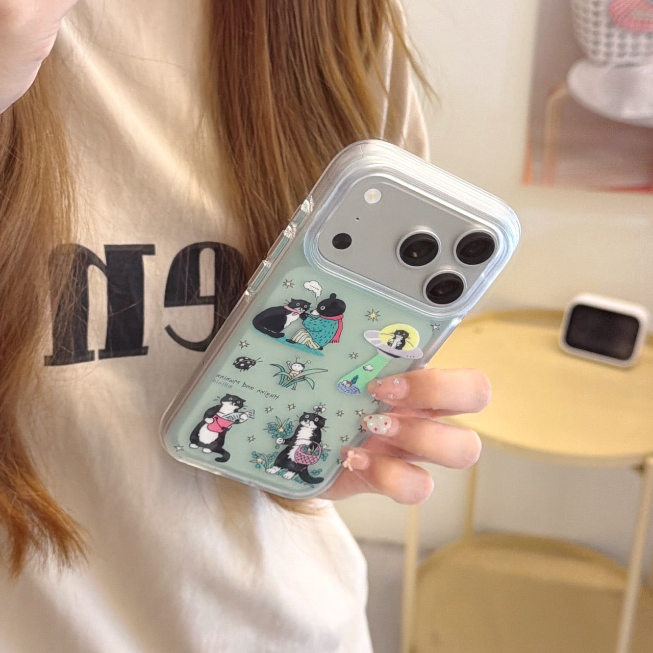 Close-up shot of a hand holding a sage green protective phone cover with functional HD makeup mirror and quirky cat graphics.