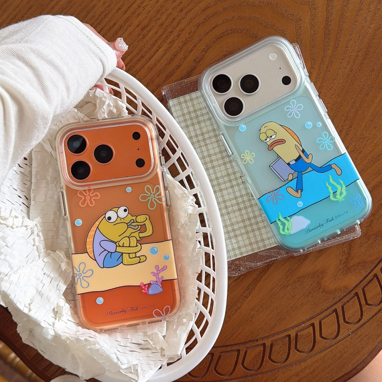 Two funny cartoon fish iPhone cases displayed on a wooden table with one placed inside a white basket.