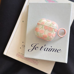 An aesthetic clear pink cherry AirPods case resting flat on a light blue Je t'aime book.
