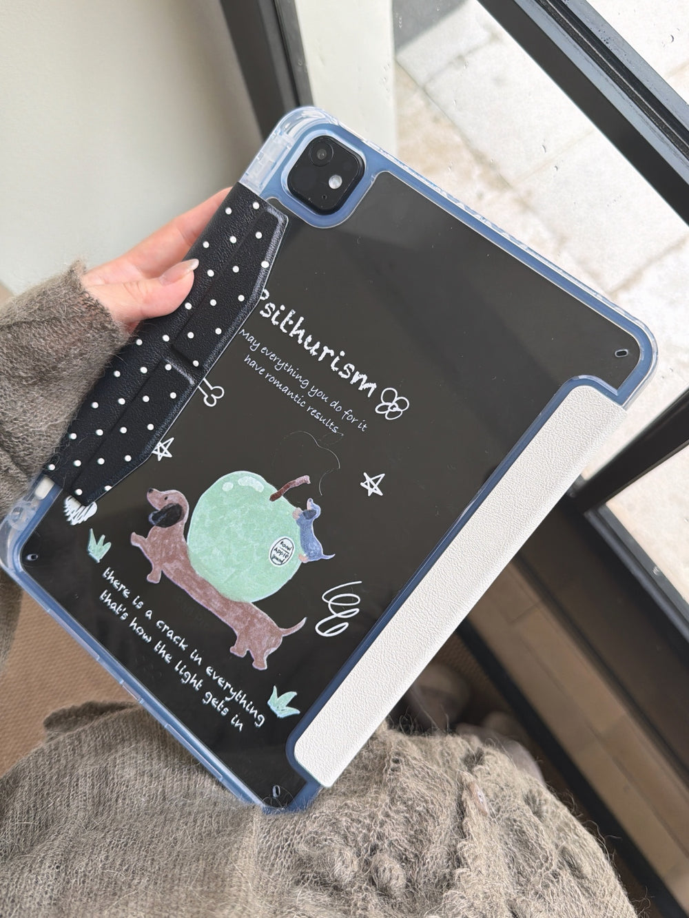 A close-up of a hand holding an aesthetic clear back ipad case, showcasing the printed cute dachshund dog graphic and "Psithurism" text on a black background, featuring a black polka-dot magnetic clasp on the side.