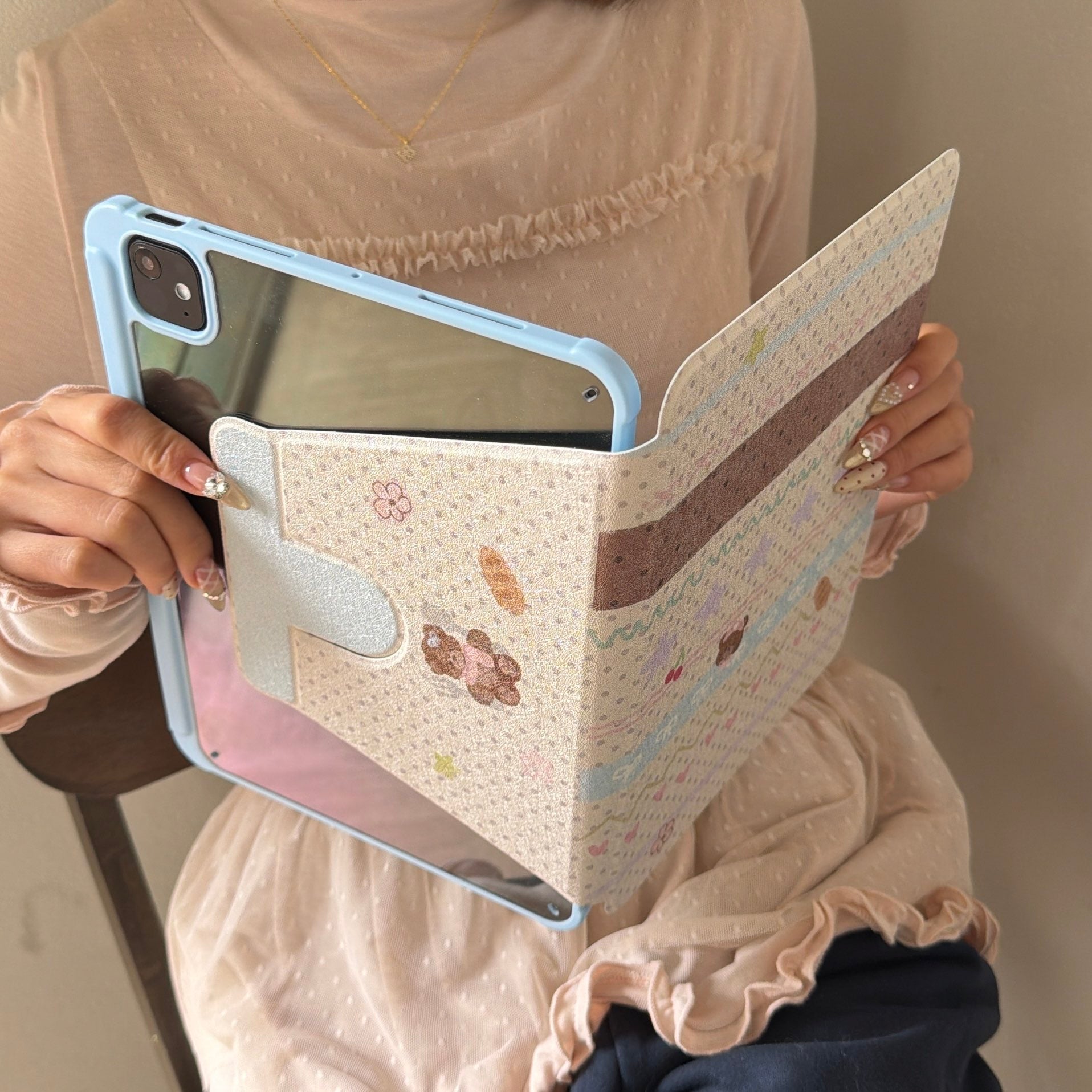 Close-up of a person holding an open pastel blue iPad case, highlighting the scratch-resistant crystal-clear back shell and the decorative vintage-style patterned front flap.