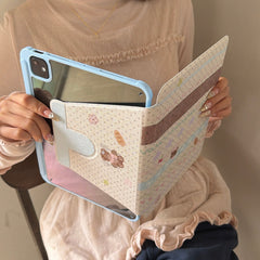 Close-up of a person holding an open pastel blue iPad case, highlighting the scratch-resistant crystal-clear back shell and the decorative vintage-style patterned front flap.