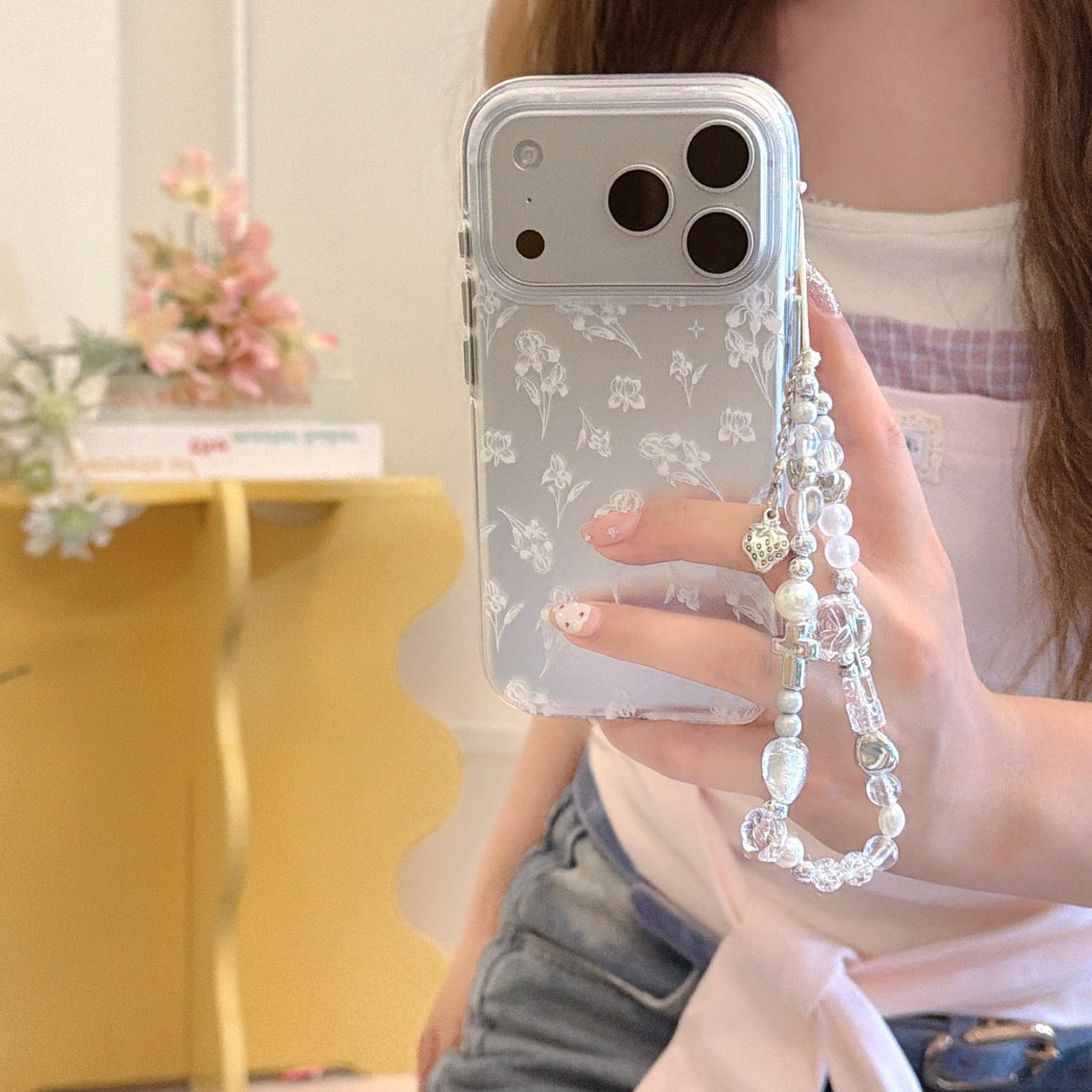 A person holding a clear floral phone case with a dangling pearl, crystal, and cross charm wristlet strap.