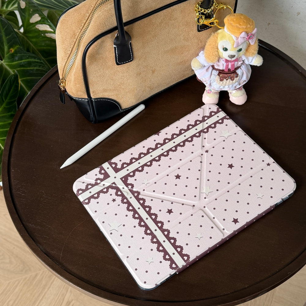 Top-down view of a closed aesthetic coquette pink iPad case on a round dark wood table next to a suede handbag, a white stylus, and a plush doll.