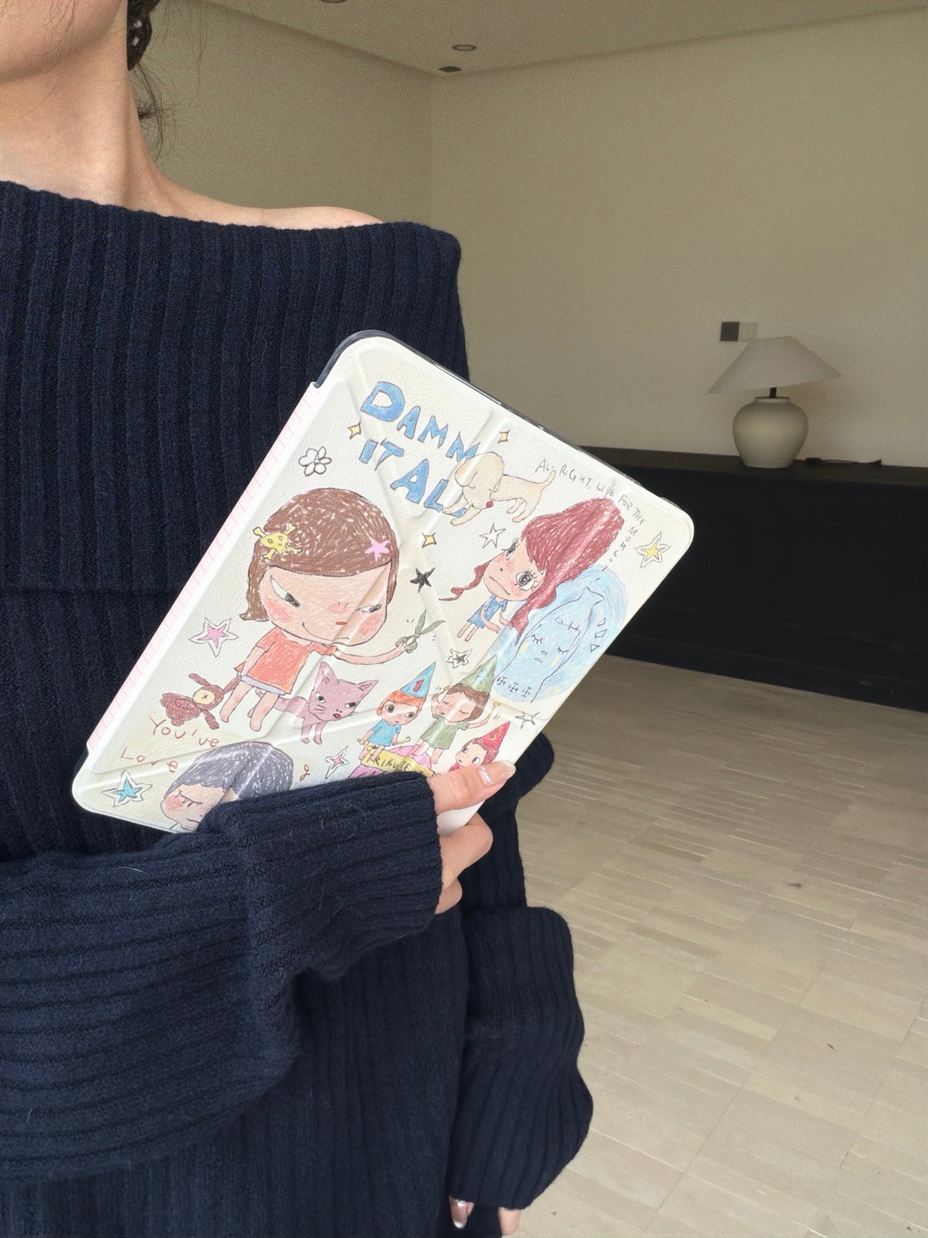 Close-up of a model wearing an off-shoulder navy blue sweater holding a cute Yoshitomo Nara art iPad cover in a minimalist indoor setting with a modern ceramic lamp.