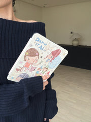 Close-up of a model wearing an off-shoulder navy blue sweater holding a cute Yoshitomo Nara art iPad cover in a minimalist indoor setting with a modern ceramic lamp.