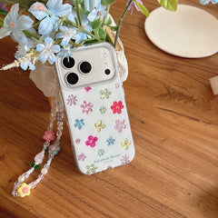 A clear aesthetic flower phone case with a beaded wristlet standing next to blue blossoms on a wooden surface.
