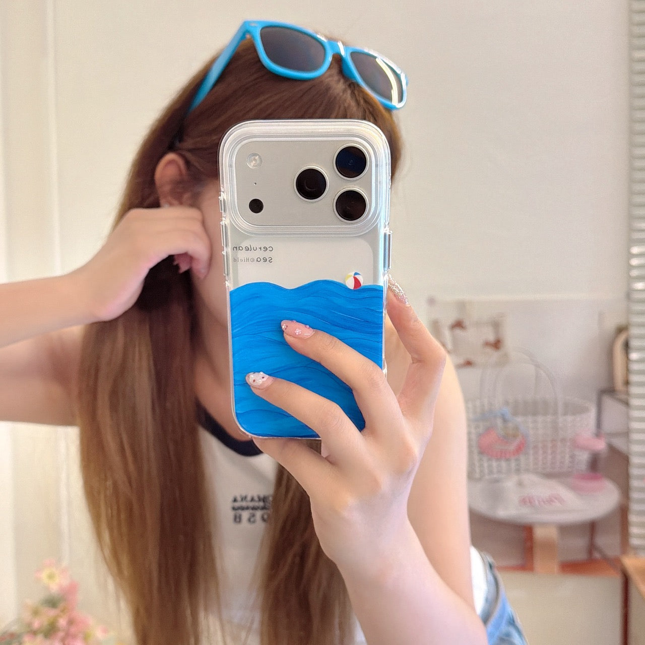 A girl taking a mirror selfie showing a clear iPhone case with a 3D textured blue ocean wave design and blue sunglasses on her head.