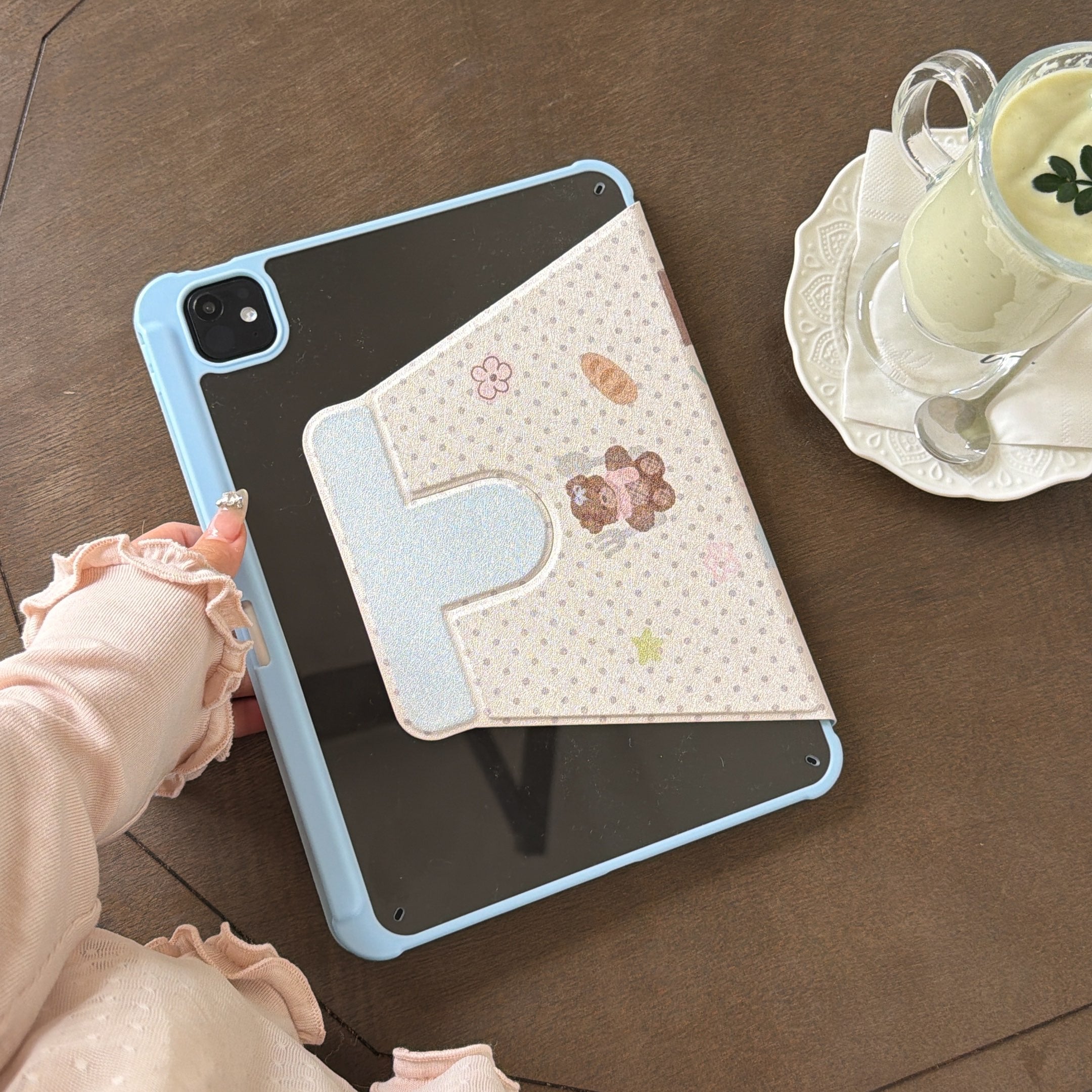 An elegant flat lay on a wooden cafe table showing an aesthetic baby blue iPad cover with its folded magnetic flap facing up, next to a glass of matcha green tea.